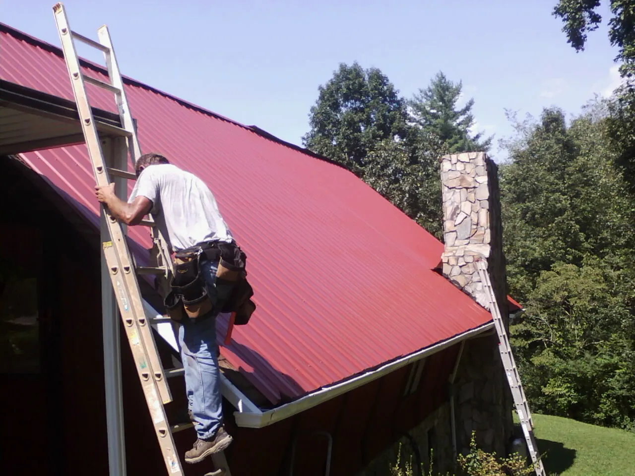 Roofer on a ladder beside a red metal roof and stone chimney — Soffit & Fascia Repair by Roofing Brothers in Peshastin